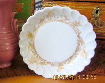 May include: A white porcelain saucer with a scalloped edge and a gold floral design. The saucer is empty.