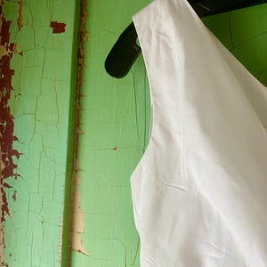 May include: A sleeveless white garment featuring a smooth fabric texture, hanging on a black plastic hanger. The bodice displays a clean, minimalist design with a simple neckline and structured shoulder strap, showcasing a crisp and bright appearance.