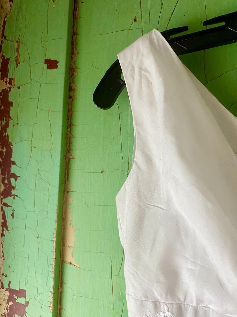 May include: A sleeveless white garment featuring a smooth fabric texture, hanging on a black plastic hanger. The bodice displays a clean, minimalist design with a simple neckline and structured shoulder strap, showcasing a crisp and bright appearance.