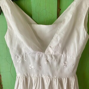 May include: A white sleeveless dress featuring a V-neckline and a fitted waist panel. The waist panel is decorated with small, delicate white floral embroidery. The skirt portion of the dress is gathered, creating soft vertical pleats.