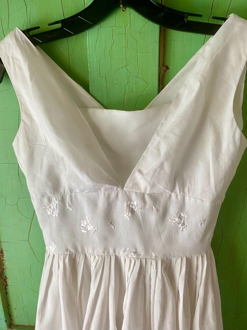 May include: A white sleeveless dress featuring a V-neckline and a fitted waist panel. The waist panel is decorated with small, delicate white floral embroidery. The skirt portion of the dress is gathered, creating soft vertical pleats.