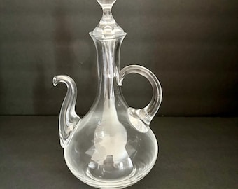 May include: A clear glass cruet with a rounded base, a slender neck, a curved handle, and a long, tapered spout. The vessel includes a matching clear glass stopper with a spherical top that fits securely into the opening of the neck.
