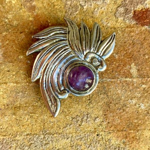 May include: A silver-toned metal brooch with a stylized feather design and a central, round, purple gemstone. The brooch is set against a textured, tan-colored surface, showcasing its intricate details and craftsmanship.