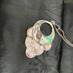 Pu&ograve; includere: Una collana d'argento con un grande ciondolo in madreperla di abalone iridescente. Il ciondolo ha la forma di un cerchio con un cerchio pi&ugrave; piccolo tagliato al centro. Il ciondolo &egrave; circondato da pezzi pi&ugrave; piccoli di madreperla di abalone.