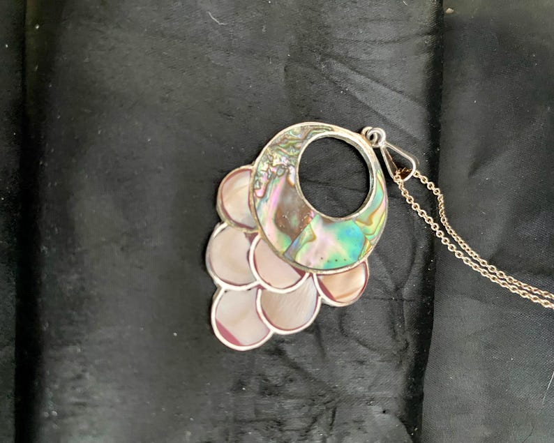Pu&ograve; includere: Una collana d'argento con un grande ciondolo in madreperla di abalone iridescente. Il ciondolo ha la forma di un cerchio con un cerchio pi&ugrave; piccolo tagliato al centro. Il ciondolo &egrave; circondato da pezzi pi&ugrave; piccoli di madreperla di abalone.
