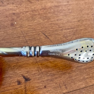 May include: Close-up of a silver-toned metal yerba mate bombilla with a perforated head and a curved handle. The word "INDUSTRIA WARFORD" is stamped on the handle. The bombilla is resting on a wooden surface.