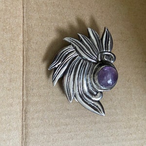 May include: A silver-toned brooch with a stylized floral design. The brooch features a central, round, purple gemstone. The metalwork has a textured, detailed appearance, with a leaf-like pattern radiating outwards from the gemstone.