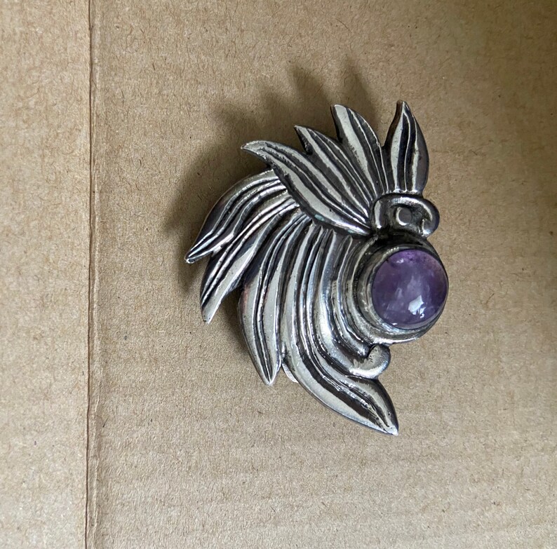 May include: A silver-toned brooch with a stylized floral design. The brooch features a central, round, purple gemstone. The metalwork has a textured, detailed appearance, with a leaf-like pattern radiating outwards from the gemstone.