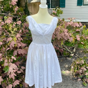 May include: A sleeveless white dress featuring a draped, crossover neckline and a fitted bodice that transitions into a full, pleated skirt. The bodice displays subtle, tonal embroidered floral details, and the dress is displayed on a mannequin.