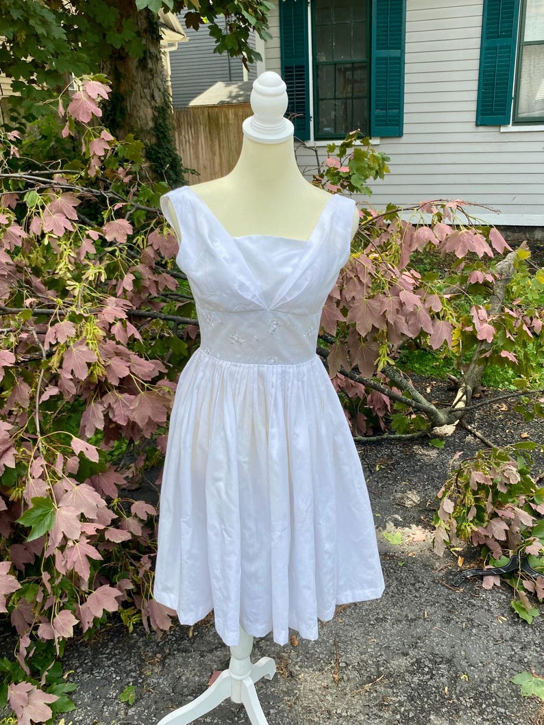 May include: A sleeveless white dress featuring a draped, crossover neckline and a fitted bodice that transitions into a full, pleated skirt. The bodice displays subtle, tonal embroidered floral details, and the dress is displayed on a mannequin.