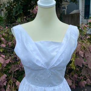 May include: A sleeveless white dress featuring a wide V-neckline with pleated fabric details at the bodice. The waist is defined by a band of white fabric adorned with subtle, tonal floral embroidery, transitioning into a full, gathered skirt.