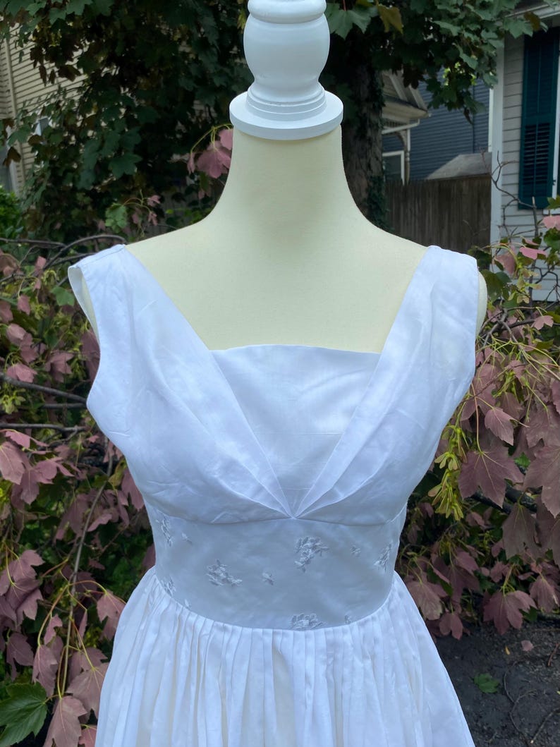 May include: A sleeveless white dress featuring a wide V-neckline with pleated fabric details at the bodice. The waist is defined by a band of white fabric adorned with subtle, tonal floral embroidery, transitioning into a full, gathered skirt.