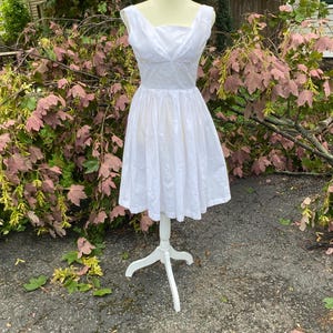 May include: A sleeveless white dress featuring a square neckline with subtle draped fabric detailing at the bodice, a fitted waistline, and a full, pleated knee-length skirt. The bodice displays delicate white floral embroidery throughout.
