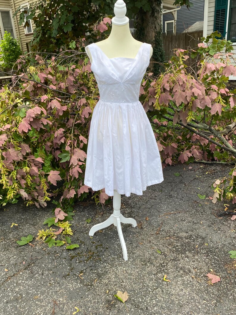 May include: A sleeveless white dress featuring a square neckline with subtle draped fabric detailing at the bodice, a fitted waistline, and a full, pleated knee-length skirt. The bodice displays delicate white floral embroidery throughout.