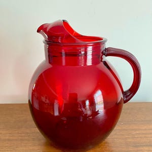 May include: A round, deep red glass pitcher featuring a smooth, bulbous body, a wide pouring spout, and a sturdy, curved handle on the side. The glass is translucent, allowing light to pass through the vibrant red color of the vessel.