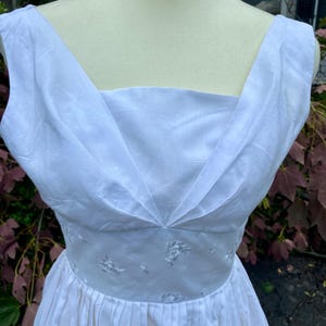 May include: A sleeveless white dress featuring a square neckline with pleated fabric detailing at the bodice. The waist is defined by a band of white fabric embroidered with small floral patterns, transitioning into a pleated skirt below the waistline.