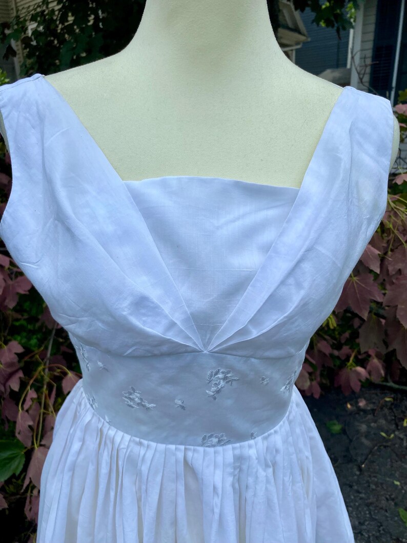 May include: A sleeveless white dress featuring a square neckline with pleated fabric detailing at the bodice. The waist is defined by a band of white fabric embroidered with small floral patterns, transitioning into a pleated skirt below the waistline.