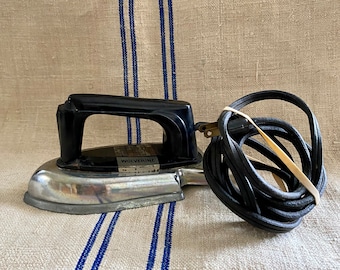 May include: A vintage electric iron with a black handle and a silver base. The iron has the word "Wolverine" printed on it. A black power cord is coiled next to the iron, secured with a rubber band. The iron is on a beige fabric with blue stripes.