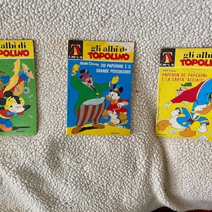 May include: Three vintage Italian comic books featuring Disney characters. The covers are brightly colored with illustrations of Mickey Mouse, Donald Duck, and Uncle Scrooge. The titles are in Italian, with the series name "Gli albi di Topolino" prominently displayed.