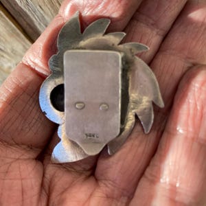 May include: A silver-toned metal brooch with a stylized floral or sunburst design. The brooch has a rectangular clasp on the back with two small holes and a stamped marking. The brooch is held in a hand.