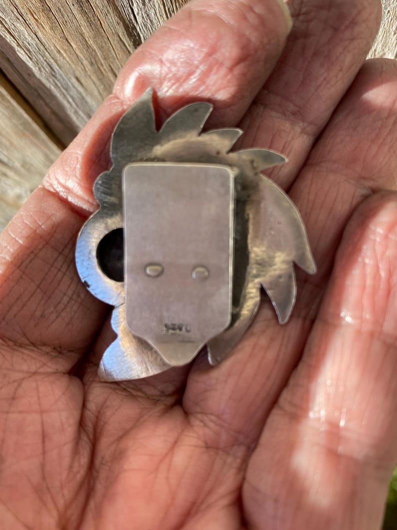 May include: A silver-toned metal brooch with a stylized floral or sunburst design. The brooch has a rectangular clasp on the back with two small holes and a stamped marking. The brooch is held in a hand.