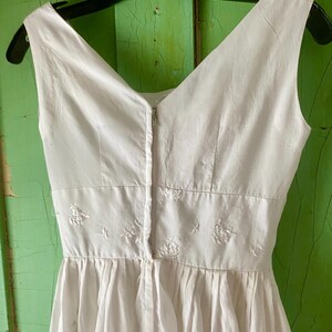 May include: A sleeveless white dress featuring a V-neckline and a fitted waist panel adorned with delicate white floral embroidery. The garment includes a central vertical zip closure running down the bodice and a gathered skirt section.
