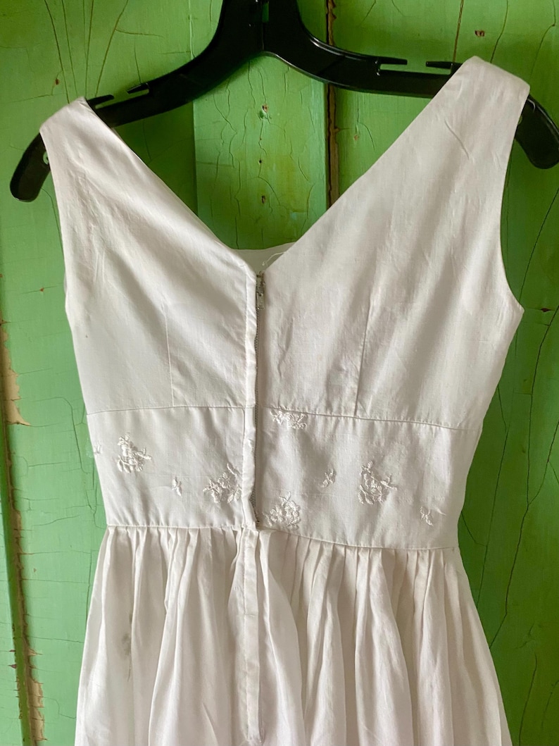 May include: A sleeveless white dress featuring a V-neckline and a fitted waist panel adorned with delicate white floral embroidery. The garment includes a central vertical zip closure running down the bodice and a gathered skirt section.