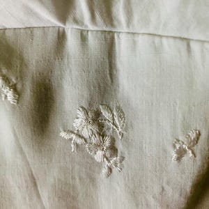 May include: A light beige fabric featuring delicate white floral embroidery. The textile displays a subtle woven texture with small, scattered stitched flower motifs and leaf patterns across the surface, creating a soft and detailed appearance.