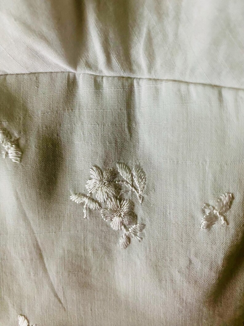 May include: A light beige fabric featuring delicate white floral embroidery. The textile displays a subtle woven texture with small, scattered stitched flower motifs and leaf patterns across the surface, creating a soft and detailed appearance.