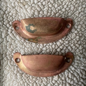 May include: Two antique copper drawer pulls with a rounded, half-circle shape. The pulls have a worn, aged finish and are slightly tarnished.