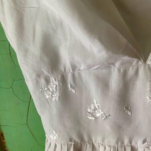 May include: A white sleeveless dress featuring a deep V-neckline and a fitted bodice. The midsection is adorned with delicate white floral embroidery. The skirt portion of the dress begins below the embroidered waistline and features soft pleats.