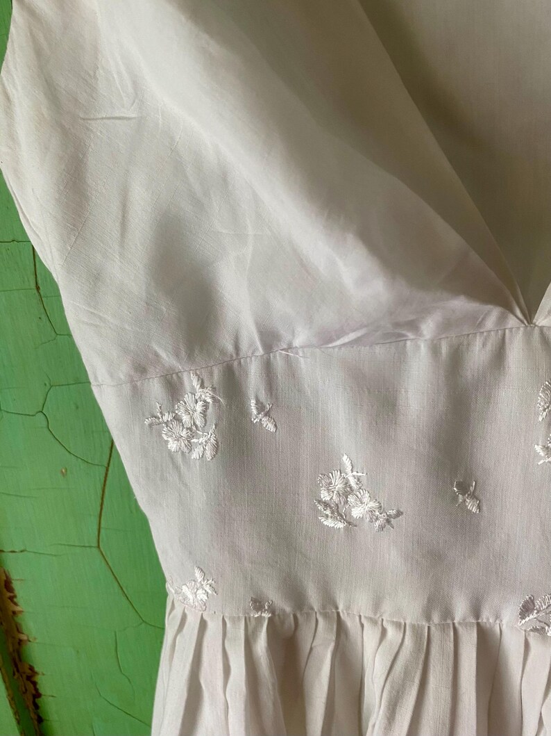 May include: A white sleeveless dress featuring a deep V-neckline and a fitted bodice. The midsection is adorned with delicate white floral embroidery. The skirt portion of the dress begins below the embroidered waistline and features soft pleats.