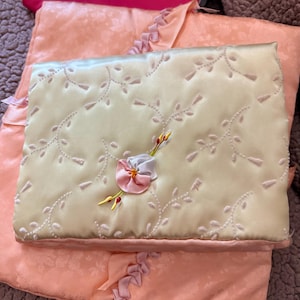 May include: A rectangular, pale yellow satin pillow featuring white embroidered vine patterns and a central three-dimensional fabric flower in shades of pink, white, and yellow. It rests atop a larger, peach-colored quilted fabric item.