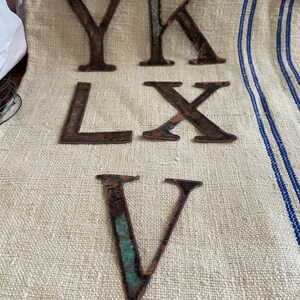 May include: Six weathered, rusty metal letters: Y, K, L, X, and V. The letters are in a dark brown color with hints of green and are arranged on a beige fabric with blue stripes. The letters appear to be vintage or antique.