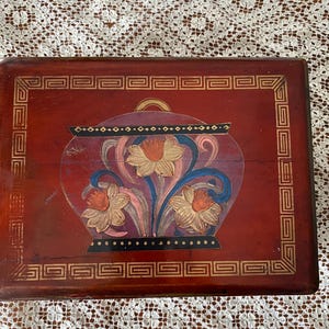 May include: A rectangular wooden box with a red background and a painted floral design. The design features a vase with three daffodils in shades of orange, pink, and blue. The box has a gold border with a Greek key pattern.