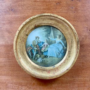 May include: A circular wall plaque featuring a central porcelain medallion with a painted scene of three figures in period clothing, set within a distressed, weathered gold-toned wooden frame with visible red underpainting and a rounded profile.