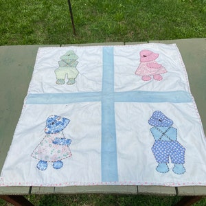 May include: A vintage baby quilt with four appliqued figures of children wearing different colored clothing. The quilt is white with blue and pink accents.