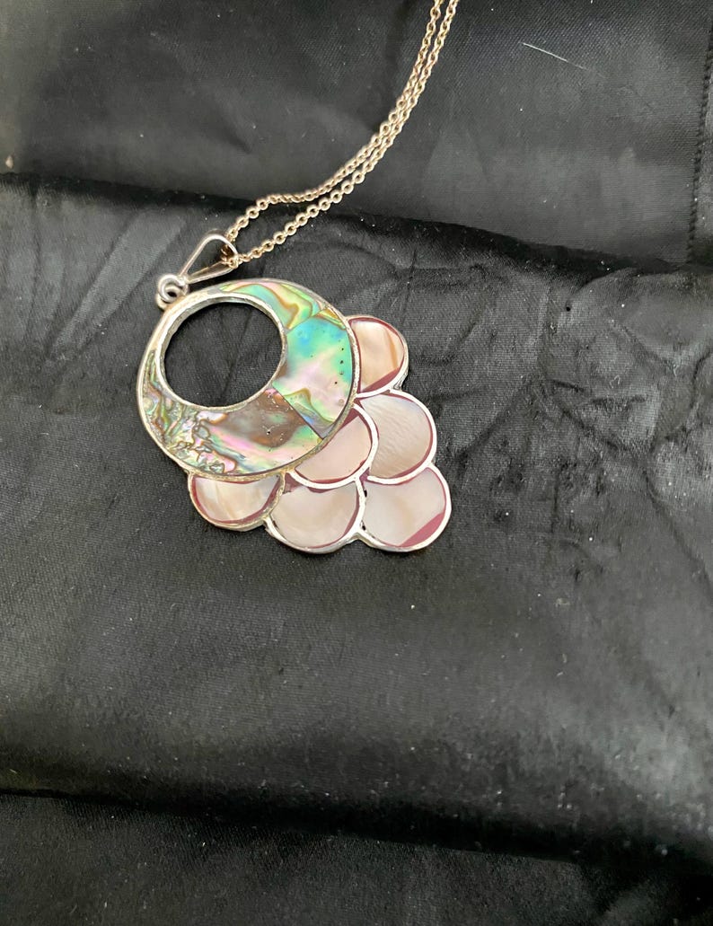 Pu&ograve; includere: Una collana con catena d'argento con un ciondolo in madreperla di abalone iridescente. Il ciondolo ha la forma di una mezzaluna con un cerchio tagliato al centro. La mezzaluna &egrave; circondata da un gruppo di cerchi pi&ugrave; piccoli in madreperla di abalone.