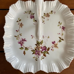 May include: A white porcelain serving dish with a scalloped edge and a handle. The dish is decorated with a floral pattern in shades of pink, purple, and green.