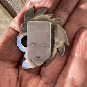 May include: A silver-toned metal brooch with a stylized floral or sunburst design. The brooch has a rectangular clasp on the back with two small holes and a stamped marking. The design features pointed petal-like shapes around a central element.