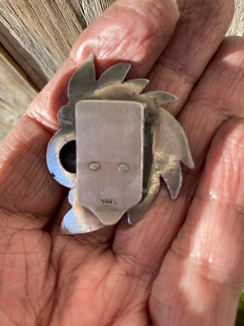 May include: A silver-toned metal brooch with a stylized floral or sunburst design. The brooch has a rectangular clasp on the back with two small holes and a stamped marking. The design features pointed petal-like shapes around a central element.
