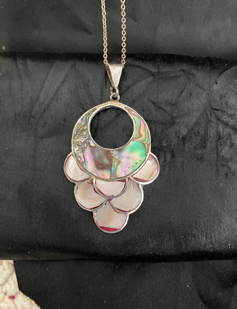 Pu&ograve; includere: Una collana d'argento con un grande ciondolo in madreperla di abalone iridescente. Il ciondolo ha la forma di un cerchio con un cerchio pi&ugrave; piccolo tagliato al centro. Il ciondolo &egrave; circondato da pezzi pi&ugrave; piccoli di madreperla di abalone.