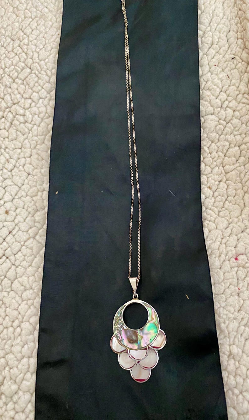 Pu&ograve; includere: Una collana con catena d'argento con un grande ciondolo in madreperla di abalone iridescente. Il ciondolo ha la forma di un cerchio con un cerchio pi&ugrave; piccolo tagliato al centro. Il ciondolo &egrave; circondato da cerchi pi&ugrave; piccoli in madreperla di abalone.