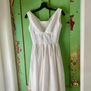 May include: A sleeveless white dress featuring a V-neckline, a fitted bodice with subtle white floral embroidery at the waist, and a gathered, knee-length skirt. The garment is displayed on a black plastic hanger against a vertical surface.