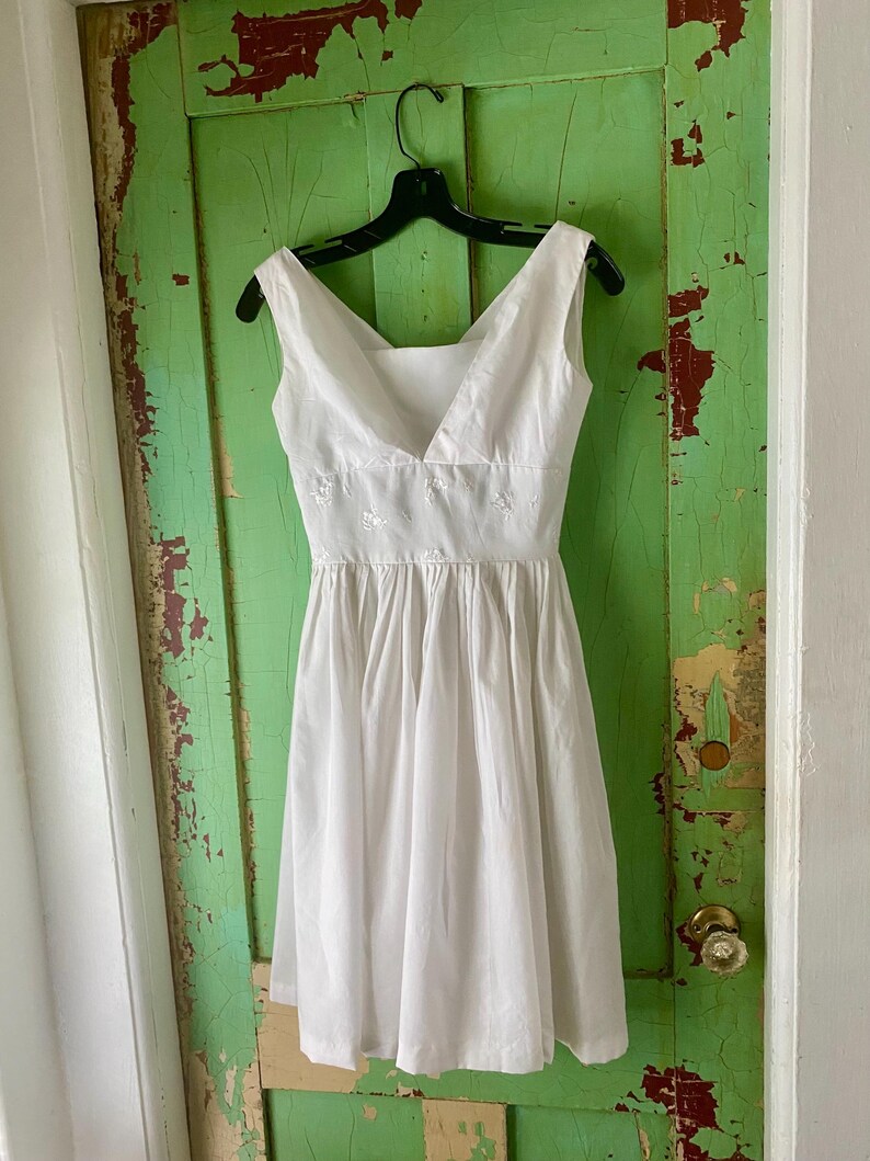 May include: A sleeveless white dress featuring a V-neckline, a fitted bodice with subtle white floral embroidery at the waist, and a gathered, knee-length skirt. The garment is displayed on a black plastic hanger against a vertical surface.