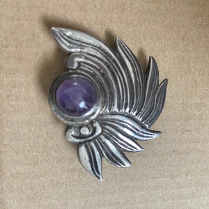 May include: A silver-toned brooch with a stylized feather design. The brooch features a round, purple gemstone at its center. The metal has a textured appearance, and the overall design is symmetrical.