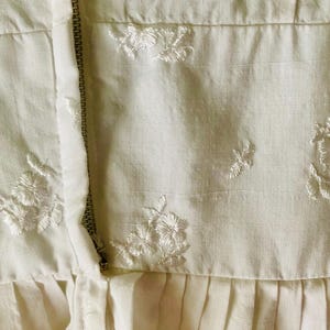 May include: A cream-coloured garment featuring a vertical metal zip closure and delicate white floral embroidery scattered across the fabric. The piece includes a horizontal seam above a pleated section, showcasing a textured weave throughout.