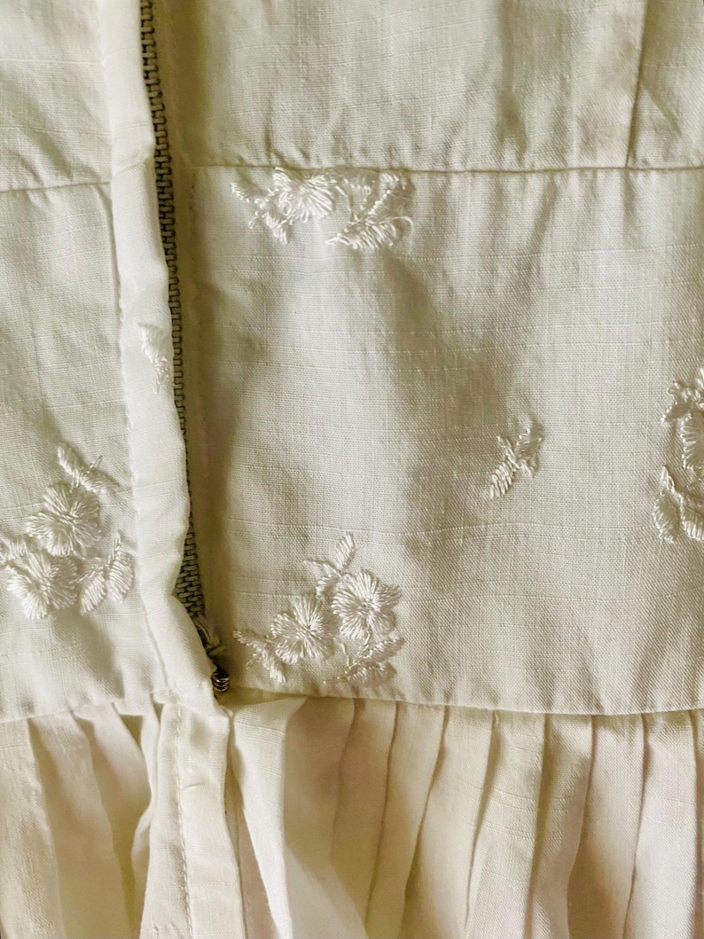 May include: A cream-coloured garment featuring a vertical metal zip closure and delicate white floral embroidery scattered across the fabric. The piece includes a horizontal seam above a pleated section, showcasing a textured weave throughout.