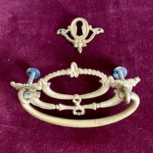 May include: Antique brass drawer pulls and keyhole cover. The drawer pull has an oval shape with a decorative design and a handle. The keyhole cover is ornate with a keyhole shape. The hardware is mounted on a burgundy background.