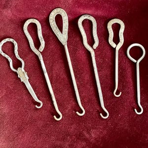 May include: A collection of vintage metal shoe button hooks and a bottle opener, arranged on a deep red velvet surface. The hooks vary in size and design, with curved handles and small hooks. Some have text on the handles.